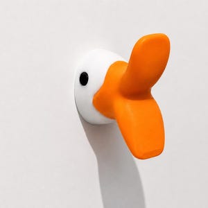 Könnte beinhalten: Ein skurriler Wandhaken in Form eines Gänsekopfes. Der Haken hat einen weißen Kopf, ein schwarzes Auge und einen leuchtend orangefarbenen Schnabel. Geeignet für Kinderzimmer oder eine verspielte, moderne Einrichtung.
