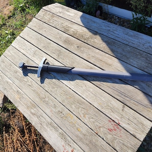 May include: A gray and black toy sword featuring a long, flat blade, a dark handle, and a silver-colored crossguard. The sword has a circular pommel at the end of the grip and a smooth, matte finish across its entire length, resting horizontally.