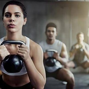 May include: A woman in a white sports bra and black shorts performs a squat while holding a black kettlebell. Other people are in the background, also exercising with kettlebells in a gym setting.