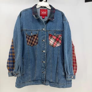 Op de afbeelding: Een blauw spijkerjack met geruite details. De jas heeft geruite zakken en mouwen in rood, geel en blauw. De kraag is ook geruit. Het heeft een knoopsluiting en het label luidt "Venezia Jeans".