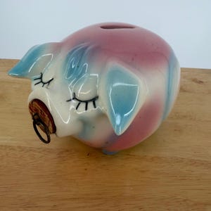 Puede incluir: Una hucha de cerámica con forma de cerdo con un acabado brillante en tonos rosa y azul claro. El cerdo presenta ojos cerrados con largas pestañas negras y un tapón de corcho en el hocico con una pequeña anilla metálica en el centro.