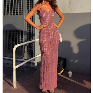 May include: A floor-length mauve crochet dress featuring a V-neckline, thin spaghetti straps, and a fitted silhouette. The garment is crafted with an open-knit texture throughout, creating a consistent pattern from the bodice down to the hemline.