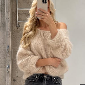 Puede incluir: Un suéter beige de punto grueso con mangas largas, un escote ancho que deja los hombros al descubierto y un acabado texturizado y esponjoso, combinado con pantalones vaqueros gris oscuro con cierre de botones y costuras visibles.