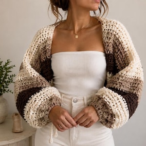 Könnte beinhalten: Gehäkeltes Bolero in Creme-, Beige- und Brauntönen. Der offene Strickjacke hat ein gestreiftes Muster und voluminöse Ärmel. Das handgefertigte Kleidungsstück hat einen entspannten, böhmischen Stil.