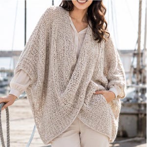 May include: A beige open-knit cardigan with a loose, draped silhouette and wide sleeves. The garment features a textured, airy weave pattern and is worn over a light-colored long-sleeved shirt and matching trousers, creating a layered look.