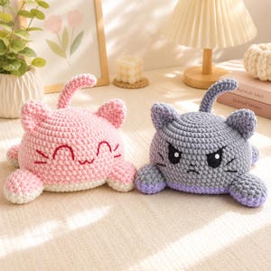 May include: Two plush cat toys made of chunky yarn. The left cat is light pink with a smiling face and closed eyes. The right cat is grey with a purple base, featuring an angry expression with furrowed brows and white-pupil eyes. Both have tails.