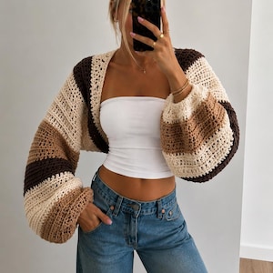 Könnte beinhalten: Ein kurzes Häkel-Shrug mit langen, voluminösen Ärmeln und horizontalen Streifen in Dunkelbraun, Hellbraun und Creme. Das Kleidungsstück hat ein offenes Vorderteil und ein lockeres, strukturiertes Strickmuster an den Ärmeln und am Rücken.