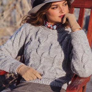 May include: A light gray cable-knit sweater with long sleeves and a crew neckline. The garment features a textured pattern of vertical cables. It is paired with a gray hat, a colorful patterned neck scarf, and tan suede gloves.