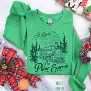 May include: A bright green crewneck sweatshirt featuring a black graphic print of a steam locomotive train traveling through a forest. The text above the train reads Believe and the text below reads The Polar Express in a script font.