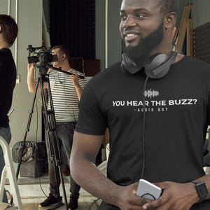 May include: A black short-sleeved t-shirt featuring a white graphic of a sound wave and the text "YOU HEAR THE BUZZ? - AUDIO GUY" printed on the chest. The shirt is worn with black over-ear headphones resting around the neck and a black wristwatch.
