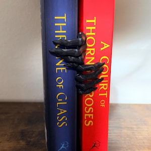 May include: A black, claw-like bookend shaped like a hand gripping the spines of two books. The blue book is titled Throne of Glass and the red book is titled A Court of Thorns and Roses. The dark, textured hand appears to be holding the books together.