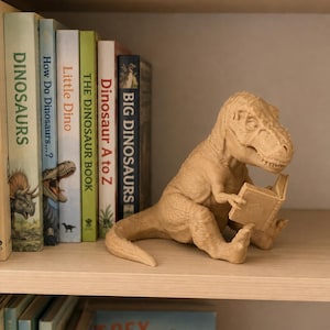 May include: A light tan, textured figurine of a Tyrannosaurus Rex sitting upright and holding an open book. The dinosaur is positioned on a wooden shelf next to a row of books with titles including Dinosaurs, How Do Dinosaurs, Little Dino, The Dinosaur Book, Dinosaur A to Z, and Big Dinosaurs.