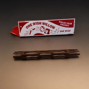 May include: A rectangular white and red box labeled ONE WISH WILLOW with the text AMAZE YOUR FRIENDS and ONE WISH, featuring a cartoon illustration of two faces. Below the box lies a brown, cylindrical, lattice-patterned plastic object.