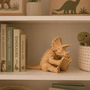 May include: A light tan triceratops figurine sitting upright and holding an open book. The dinosaur has detailed skin texture, a large frill, and three horns. It is positioned on a white shelf next to a row of books titled How Do Dinosaurs, Good Night, Dinosaur, Dinosaurs, and Dinosaur ABC.