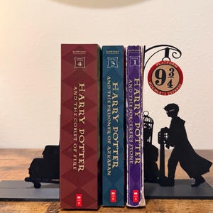 May include: Three Harry Potter books held by black metal bookends. The books are titled Harry Potter and the Goblet of Fire, Harry Potter and the Prisoner of Azkaban, and Harry Potter and the Sorcerer's Stone. The bookends feature a silhouette of a person and a Platform 9 3/4 sign.