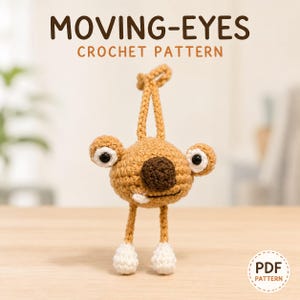May include: A tan crochet creature with a dark brown nose, two white eyes with black pupils, and two white feet. The figure has a loop on top for hanging. Text above reads MOVING-EYES CROCHET PATTERN, and a circular badge says PDF PATTERN.