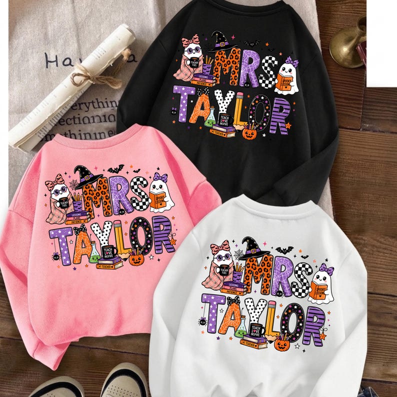 K&ouml;nnte beinhalten: Drei Rundhals-Sweatshirts in Schwarz, Pink und Wei&szlig;, jeweils mit einem Grafikdruck mit der Aufschrift MRS TAYLOR. Das Design zeigt Cartoon-Geister, einen Hexenhut, B&uuml;cher, einen K&uuml;rbis und einen Bleistift, verziert mit bunten Mustern.
