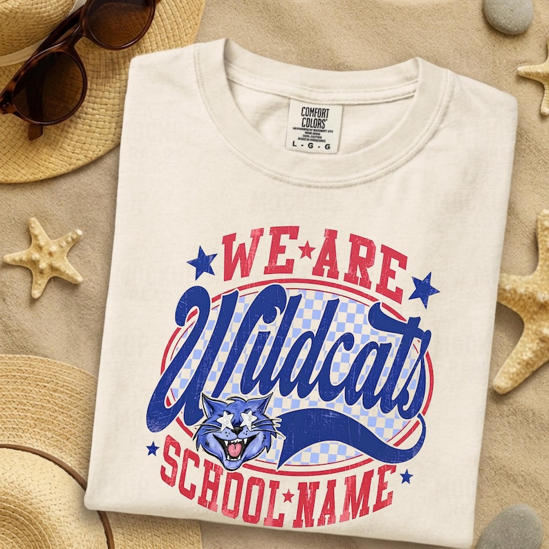 K&ouml;nnte beinhalten: Ein cremefarbenes T-Shirt mit einem Aufdruck, der den Text WE ARE WILDCATS SCHOOL NAME in Rot und Blau zeigt. Das Design enth&auml;lt ein blaues Katzengesicht mit sternf&ouml;rmigen Augen, einen karierten Hintergrund sowie rote und blaue Sterne.