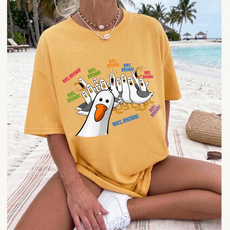 May include: A mustard yellow short-sleeve t-shirt featuring a graphic print of multiple white and grey seagulls with orange beaks. The seagulls are depicted with open mouths, and the text "MRS. BROWN!" is printed in various colors around them.