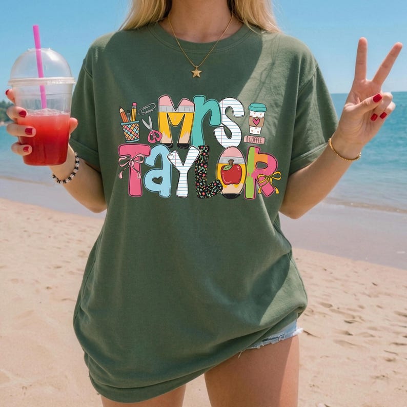 May include: An olive green short-sleeve t-shirt featuring a colorful graphic print with the text "Mrs. Taylor" in a playful font. The design includes illustrations of school supplies like pencils, scissors, a notebook, an apple, and a coffee cup.