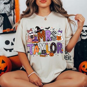 K&ouml;nnte beinhalten: Ein beiges Kurzarm-T-Shirt mit einer bunten Halloween-Grafik und dem Text MRS TAYLOR. Das Design zeigt Geister, einen Hexenhut, B&uuml;cher, einen Bleistift, einen K&uuml;rbis und Spinnennetze in orangefarbenen, violetten und schwarzen Mustern.