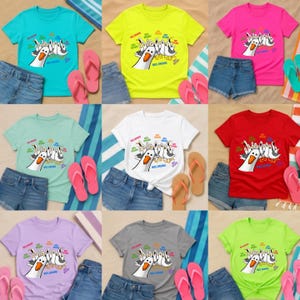 May include: A collection of nine short-sleeve t-shirts in various colors, each featuring a graphic print of multiple seagulls and the text "MRS. BROWN" in colorful lettering. The shirts are displayed alongside denim shorts and flip-flops.
