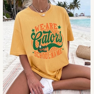 K&ouml;nnte beinhalten: Ein senfgelbes Kurzarm-T-Shirt mit einem Grafikdruck, der den Text "WE ARE Gators SCHOOL NAME" in Gr&uuml;n und Orange zeigt. Das Design umfasst einen karierten ovalen Hintergrund, gr&uuml;ne Sterne und eine Illustration eines Alligatorkopfes.