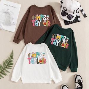 May include: Three long-sleeve crewneck sweatshirts in brown, dark green, and white. Each features a colorful graphic print with the text "Mrs. Taylor" surrounded by school-themed icons like pencils, scissors, a coffee cup, and a red apple.