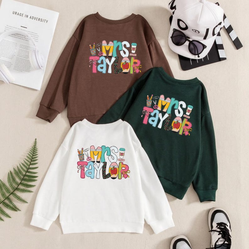 May include: Three long-sleeve crewneck sweatshirts in brown, dark green, and white. Each features a colorful graphic print with the text "Mrs. Taylor" surrounded by school-themed icons like pencils, scissors, a coffee cup, and a red apple.