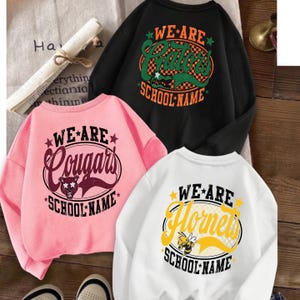 K&ouml;nnte beinhalten: Drei Rundhals-Sweatshirts in Schwarz, Pink und Wei&szlig;. Jedes zeigt eine kreisf&ouml;rmige Grafik mit dem Text "WE ARE" oben, einer Maskottchen-Illustration und "SCHOOL NAME" unten. Das schwarze Shirt zeigt "Gators", das pinke "Cougars" und das wei&szlig;e "Hornets".