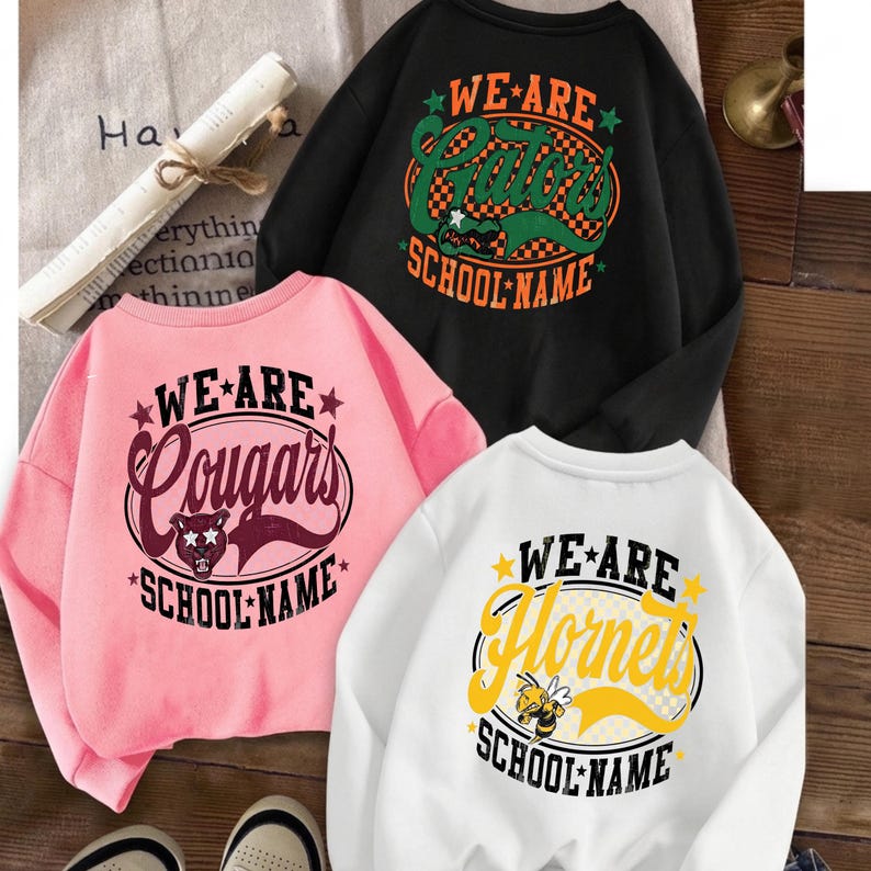 K&ouml;nnte beinhalten: Drei Rundhals-Sweatshirts in Schwarz, Pink und Wei&szlig;. Jedes zeigt eine kreisf&ouml;rmige Grafik mit dem Text "WE ARE" oben, einer Maskottchen-Illustration und "SCHOOL NAME" unten. Das schwarze Shirt zeigt "Gators", das pinke "Cougars" und das wei&szlig;e "Hornets".