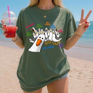 May include: An olive green short-sleeve t-shirt featuring a graphic print of multiple white seagulls with orange beaks. The seagulls are depicted with open mouths and the text "MRS. BROWN!" is printed in various colors around the birds.