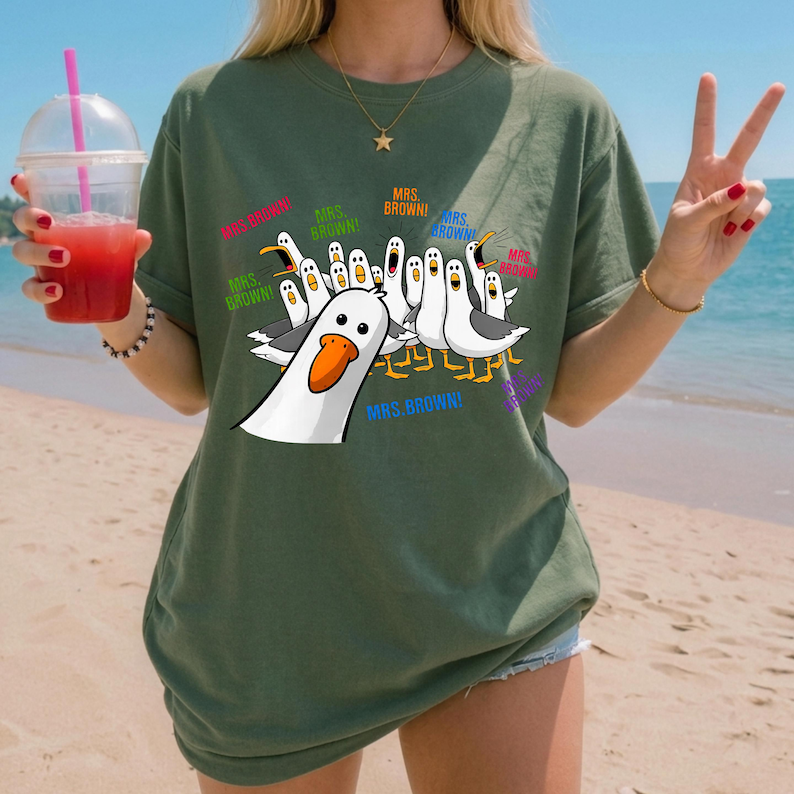 May include: An olive green short-sleeve t-shirt featuring a graphic print of multiple white seagulls with orange beaks. The seagulls are depicted with open mouths and the text "MRS. BROWN!" is printed in various colors around the birds.