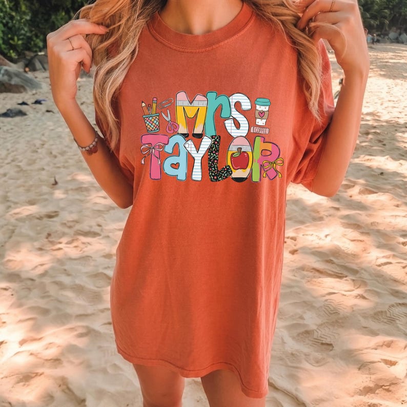 May include: A rust-colored short-sleeve t-shirt featuring a colorful graphic print that reads Mrs. Taylor. The design incorporates school-themed illustrations including pencils, scissors, a coffee cup, and a red apple, all rendered in various patterns.