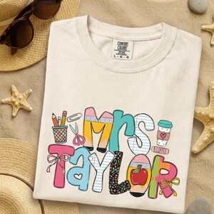 May include: A cream-colored t-shirt with a graphic print featuring the text Mrs. Taylor. The design incorporates school-themed illustrations including pencils, a cup, scissors, a paperclip, a coffee cup with a heart, and a red apple.