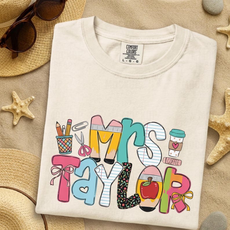 May include: A cream-colored t-shirt with a graphic print featuring the text Mrs. Taylor. The design incorporates school-themed illustrations including pencils, a cup, scissors, a paperclip, a coffee cup with a heart, and a red apple.