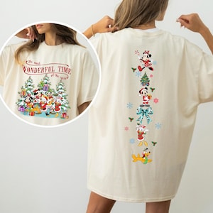 May include: An off-white t-shirt featuring a Christmas-themed graphic. The front displays Disney characters with snow-covered trees and the text "The most WONDERFUL TIME of the year." The back shows a vertical arrangement of Disney characters, a tree, and a bow.