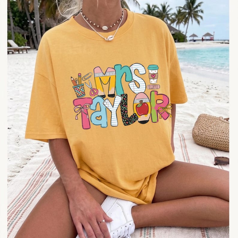 May include: A mustard yellow short-sleeve t-shirt featuring a colorful graphic print that reads "Mrs. Taylor" in large, stylized letters. The design includes illustrations of school supplies like pencils, scissors, an apple, and a coffee cup.