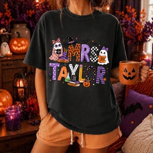 K&ouml;nnte beinhalten: Ein schwarzes Kurzarm-T-Shirt mit einem Grafikdruck und dem Text MRS TAYLOR. Das Design zeigt Cartoon-Geister, B&uuml;cherstapel, einen Hexenhut, einen Bleistift und einen K&uuml;rbis, die mit Leoparden-, Karomustern und Streifen verziert sind.