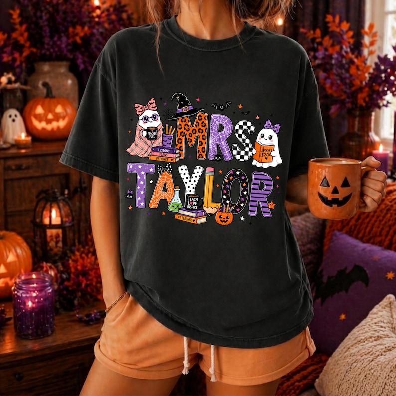 K&ouml;nnte beinhalten: Ein schwarzes Kurzarm-T-Shirt mit einem Grafikdruck und dem Text MRS TAYLOR. Das Design zeigt Cartoon-Geister, B&uuml;cherstapel, einen Hexenhut, einen Bleistift und einen K&uuml;rbis, die mit Leoparden-, Karomustern und Streifen verziert sind.