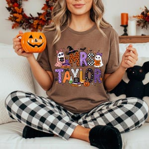 K&ouml;nnte beinhalten: Ein braunes Kurzarm-T-Shirt mit einem Grafikdruck mit dem Text MRS TAYLOR, umgeben von Illustrationen von Geistern, einem Hexenhut, B&uuml;chern, einem Bleistift und einem K&uuml;rbis in den Farben Orange, Lila und Schwarz.