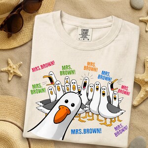 May include: A cream-colored t-shirt featuring a graphic of multiple cartoon seagulls with orange beaks and grey wings. The seagulls are shouting the text "MRS. BROWN!" in various colors including pink, green, orange, blue, and purple.