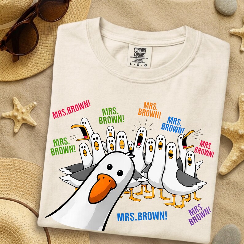 May include: A cream-colored t-shirt featuring a graphic of multiple cartoon seagulls with orange beaks and grey wings. The seagulls are shouting the text "MRS. BROWN!" in various colors including pink, green, orange, blue, and purple.