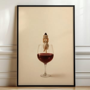 Könnte beinhalten: Ein gerahmter Wandkunstdruck, der eine kleine Figur in einem hellen Kleid zeigt, die auf dem Rand eines großen Glases mit tiefrotem Wein sitzt. Die Figur hält ein offenes Buch vor ihr Gesicht. Der Hintergrund ist in einem einheitlichen Cremeton.