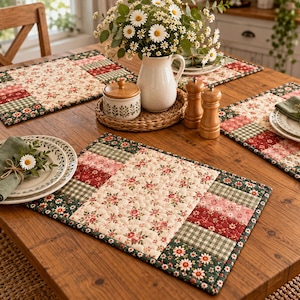 May include: Rectangular quilted placemats featuring a central cream panel with a small red and green floral print, bordered by patchwork strips of red floral, green gingham, and dark green daisy patterns, finished with a dark green binding edge.