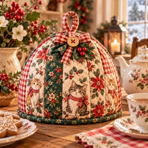 May include: A quilted tea cozy featuring a festive holiday pattern with kittens, red poinsettias, and green holly leaves. The fabric has alternating panels of red and white gingham and floral prints, topped with a fabric loop and a wooden button.