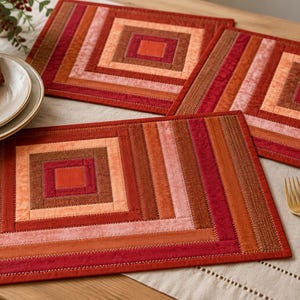 May include: Three rectangular fabric placemats featuring a quilted log cabin pattern in shades of red, orange, pink, and brown. Each placemat is constructed from stitched fabric strips arranged in concentric squares, creating a geometric design.