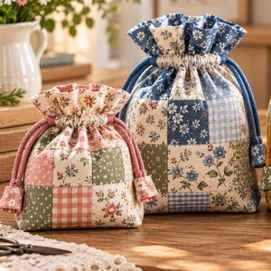 May include: Two fabric drawstring pouches featuring a patchwork design. The smaller pouch has pink, green, and cream floral and gingham patterns with pink cords. The larger pouch has blue, white, and cream floral and gingham patterns with blue cords.