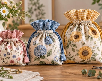 May include: Three fabric drawstring pouches of varying sizes. The smallest has a pink floral print, the medium features blue hydrangea blossoms, and the largest displays yellow sunflowers with a yellow and white gingham top trim.