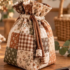 May include: A drawstring pouch crafted from a patchwork of fabric squares featuring various floral prints and a brown and white gingham pattern. The bag is cinched at the top with thick brown braided cords ending in fabric-covered beads.